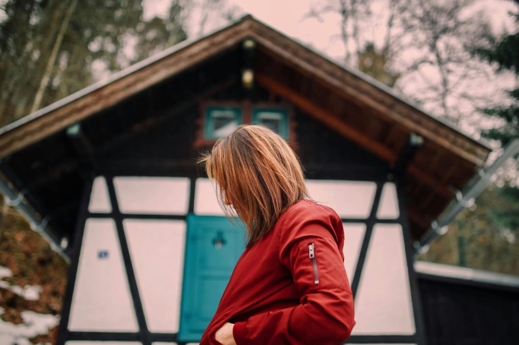 A woman in a red jacket stands outdoors near a rustic timber house, creating a cozy autumn scene.