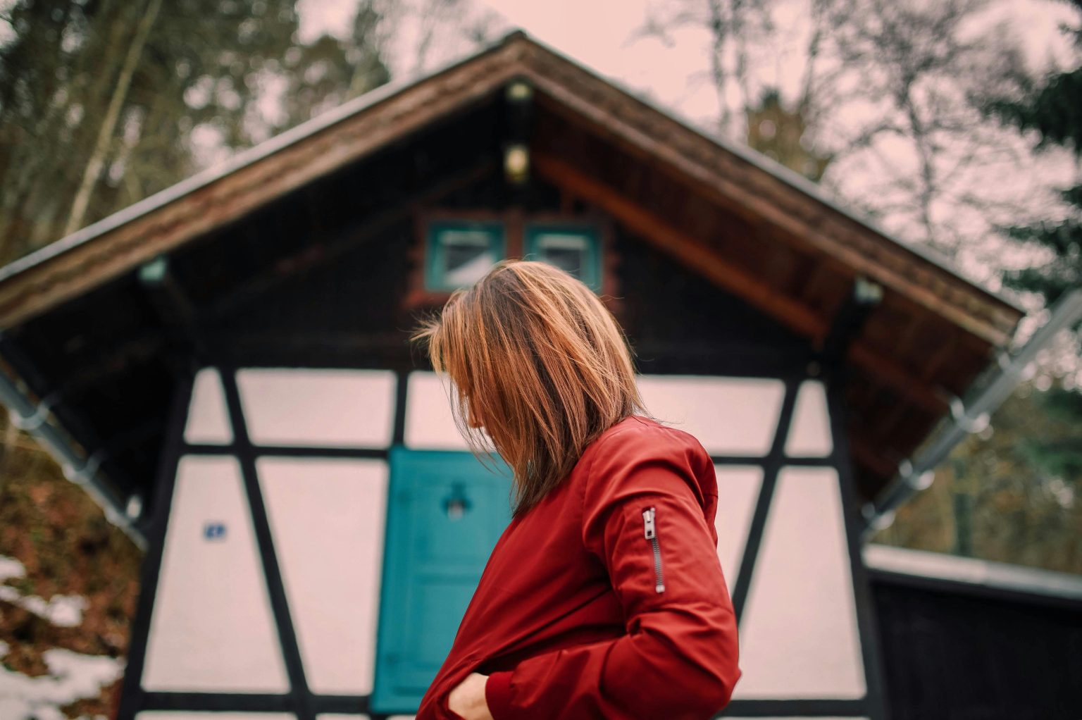A woman in a red jacket stands outdoors near a rustic timber house, creating a cozy autumn scene.
